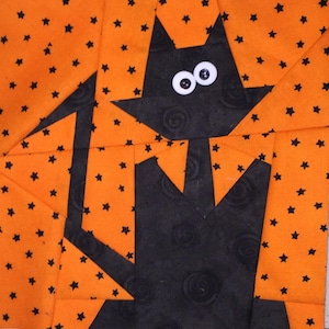 May include: A fabric quilt block featuring a black cat silhouette with large white googly eyes against a bright orange background patterned with small black stars. The cat is constructed from black fabric with a subtle swirl texture pattern.