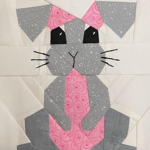 Puede incluir: Un bloque de tela con un conejito gris con un lazo y una corbata rosas. El conejito tiene ojos negros y bigotes de hilo negro. El fondo es blanco con líneas diagonales, creando un diseño de patchwork.
