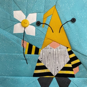 Könnte beinhalten: Ein gelber und schwarz gestreifter Zwerg mit einem gelben Hut und einer weißen Blume mit gelber Mitte. Der Zwerg hat schwarze Augen und ein rosafarbenes Gesicht.