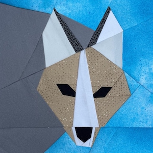 May include: A fabric quilt block featuring a stylized fox head design. The fox is composed of tan speckled fabric, white accents, and black eyes and nose. It is set against a background of solid dark gray and mottled bright blue fabric pieces.