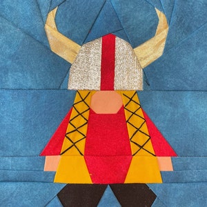 Peut inclure: Une application en tissu rouge et jaune représentant un Viking avec un casque argenté et des cornes dorées. Le Viking porte un gilet jaune avec des coutures noires.