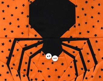 May include: A black spider with white button eyes is appliqued on an orange fabric background with black stars. The spider has eight legs and a large body.