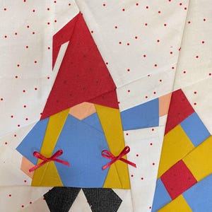 May include: A patchwork quilt block featuring a gnome with a red hat, blue shirt, yellow pants, and red bows. The background is white with red polka dots.