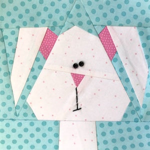 Puede incluir: Un bloque de tela con un diseño de cara de conejo. La cara del conejo es blanca con orejas rosas con lunares y una nariz rosa. Dos pequeños botones negros forman los ojos, sobre un fondo azul claro con lunares blancos. El diseño es de estilo patchwork.