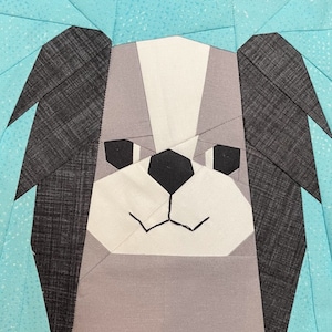 Op de afbeelding: Een close-up van een grijze en witte stoffen hond met zwarte oren. De hond heeft een gestikte neus en mond.