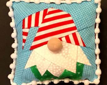 May include: A handmade gnome ornament with a red and white striped hat, a white beard, and a green and white patterned body. The gnome is sewn onto a blue and white checkered fabric square with a white pom-pom trim.