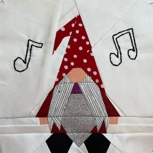 May include: A white fabric square with a red and white polka dot gnome applique. The gnome is wearing a gray and white striped shirt and a purple vest. There are two black musical notes stitched on the fabric.