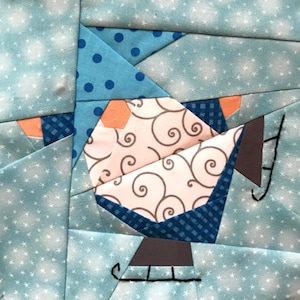 Puede incluir: Un bloque de colcha de retazos con un diseño de copo de nieve azul y blanco con un pequeño muñeco de nieve naranja y blanco que sostiene un trineo marrón y gris.