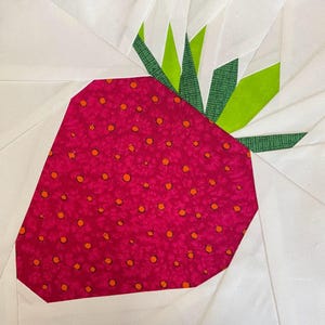 Peut inclure: Un bloc de courtepointe en tissu représentant un motif central de fraise rose foncé avec de petits points orange. La fraise possède des feuilles vertes texturées au sommet, sur un fond de pièces de tissu blanc en motif géométrique.