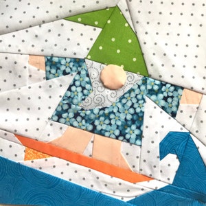 Peut inclure: Un bloc de courtepointe en patchwork représentant un gnome portant un chapeau vert et une chemise à fleurs bleues, sur une planche de surf sur une vague bleue. L'arrière-plan est blanc avec des pois gris.