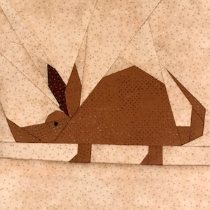 Può includere: Un blocco patchwork in tessuto marrone e beige con un armadillo stilizzato. L'armadillo è realizzato in tessuto marrone con una trama a pois, e lo sfondo è un tessuto beige chiaro con piccoli punti bianchi. Il design è geometrico e sembra cucito a mano.