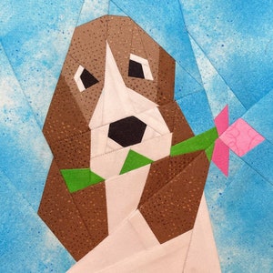 May include: A brown and white dog with a pink flower in its mouth. The dog is made of fabric and is part of a quilt.