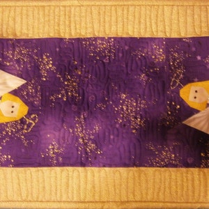 May include: A purple table runner with a gold border features two white angels with yellow hair and gold halos. The angels are holding a gold star and a gold bell. The background is a purple fabric with gold glitter.