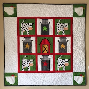 Op de afbeelding: Een wit quilt met een rood, groen en grijs patchwork ontwerp. De quilt heeft een schuur, koeien, melkbussen en kippen.