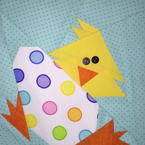 May include: A yellow chick peeking out of a white egg with colorful polka dots. The chick has black eyes and an orange beak. The background is a light blue fabric with white polka dots.