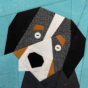 Op de afbeelding: Een patchwork quiltblok met het gezicht van een hond. De hond is zwart, wit en bruin met witte knopen als ogen. De achtergrond is een lichtblauwe, gespikkelde stof.