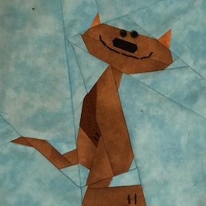 Puede incluir: Un gato marrón hecho de cuadrados de tela, con ojos negros y una boca negra, está sentado sobre un fondo azul.