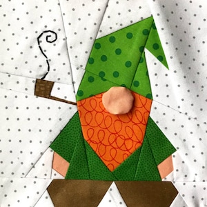 Puede incluir: Un gnomo verde con barba marrón y una camisa con estampado rojo y naranja. El gnomo lleva un gorro verde con lunares blancos y sostiene una pipa de la que sale humo.