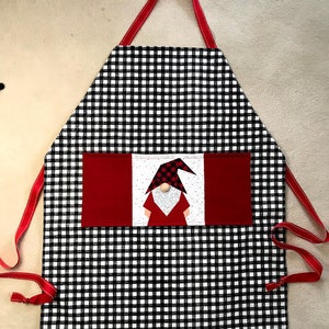 Może przedstawiać: Czarno-biały fartuch w kratkę z czerwoną kieszenią ozdobioną aplikacją filcowego gnoma. Gnom ma czerwony kapelusz w białe kropki i białą brodę.