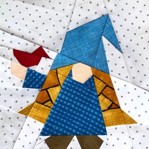 Peut inclure: Un bloc de courtepointe en patchwork représentant un gnome avec un chapeau bleu, une veste marron, une chemise bleue et des chaussures rouges. Le gnome est debout sur un fond blanc à pois gris.