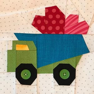 May include: A green toy truck with a blue bed and black wheels. The truck is carrying a red and pink gift with white polka dots and pink stripes.