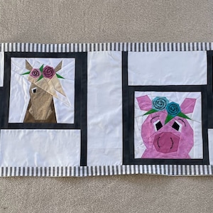 May include: A white fabric table runner with black stripes and a patchwork design featuring five animal faces with floral headbands. The animals include a cat, a horse, a pig, and two rabbits.