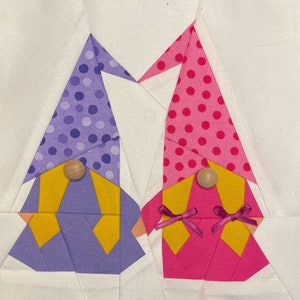 Pode incluir: Dois gnomos de tecido, um roxo com detalhes amarelos e outro rosa com detalhes amarelos, são costurados juntos. Ambos os gnomos têm bolinhas brancas nos seus chapéus e narizes de madeira.