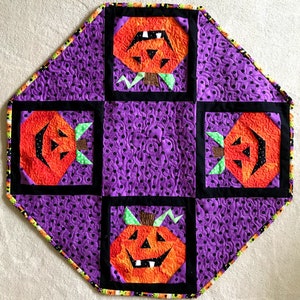 May include: A purple quilted table runner with a black and white polka dot pattern. The runner features four orange and black jack-o'-lantern appliques.