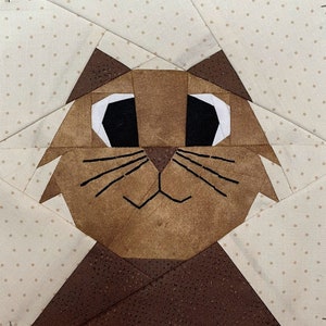 May include: A fabric block featuring a cat face design. The cat's face is brown with black whiskers and white and black eyes. The background is a cream color with small brown polka dots. The cat's ears are a darker shade of brown.