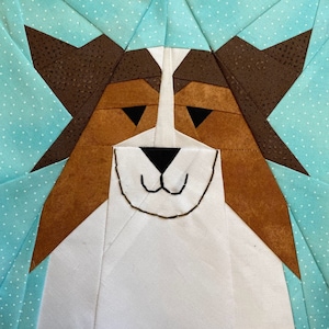 May include: A patchwork quilt block featuring a dog's face. The dog has brown ears, brown and orange fur, and a white face with black eyes and a stitched-on nose and mouth.