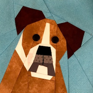 Puede incluir: Una obra de arte textil que representa la cara de un perro estilizado. El perro tiene pelaje marrón y blanco, orejas marrones oscuras y ojos negros de botón. El fondo es una tela azul claro con un patrón sutil. La nariz del perro es negra y gris.