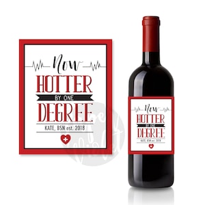 May include: Red wine bottle with a white label that says "Now Hotter By One Degree Kate, BSN est. 2018" with a red heart and a plus sign.
