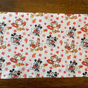 May include: White cotton kitchen towel with a repeating pattern of Mickey Mouse and Minnie Mouse holding hands with Donald Duck and Daisy Duck. The pattern is in red, black, and orange.