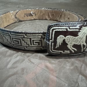 May include: A brown leather belt with a white horse design on the buckle. The belt has a geometric pattern embroidered in white thread.