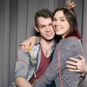 May include: A couple poses for a photo, the woman wearing a crown headband and the man with a necklace. They are both smiling and looking at the camera.