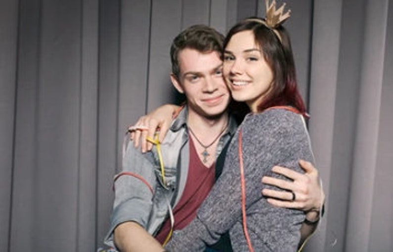 May include: A couple poses for a photo, the woman wearing a crown headband and the man with a necklace. They are both smiling and looking at the camera.