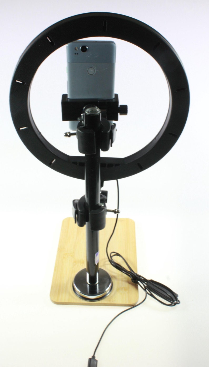 May include: A black ring light with a phone holder attached to a black metal stand. The stand has a wooden base and a black cord.