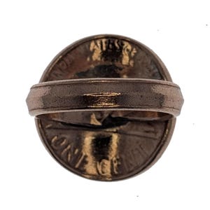 Puede incluir: Un anillo de color cobre con una moneda estadounidense de un centavo incrustada en la parte superior. La moneda presenta el perfil de Abraham Lincoln y las palabras "LIBERTY" y "ONE CENT".