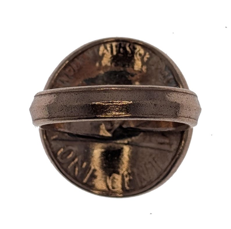 Puede incluir: Un anillo de color cobre con una moneda estadounidense de un centavo incrustada en la parte superior. La moneda presenta el perfil de Abraham Lincoln y las palabras "LIBERTY" y "ONE CENT".