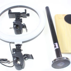 May include: A white ring light with a black adjustable phone holder and a black stand with a magnetic base. The stand is attached to a wooden cutting board.
