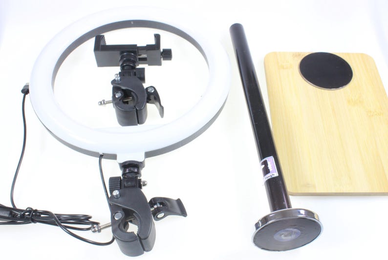 May include: A white ring light with a black adjustable phone holder and a black stand with a magnetic base. The stand is attached to a wooden cutting board.
