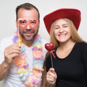 May include: A couple wearing festive accessories for a Valentine's Day celebration. The man is wearing a lei and heart-shaped glasses, while the woman is wearing a red cowboy hat and holding a red heart on a stick.