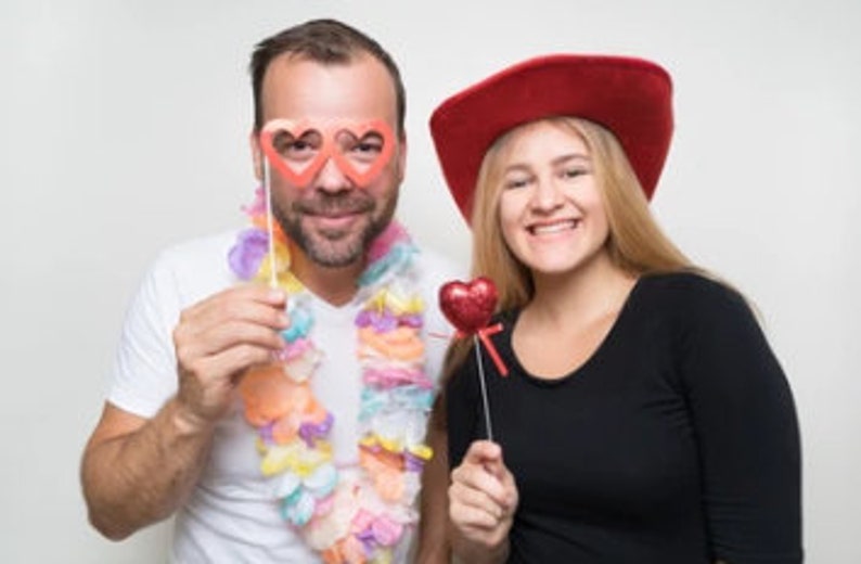 May include: A couple wearing festive accessories for a Valentine's Day celebration. The man is wearing a lei and heart-shaped glasses, while the woman is wearing a red cowboy hat and holding a red heart on a stick.