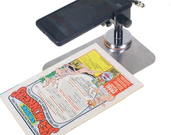 Puede incluir: Un soporte de teléfono negro con una base plateada y una placa magnética. El soporte sostiene un teléfono inteligente negro sobre un cómic con el texto "Super Sea Monkeys" y "Just add water - create live instant pets".