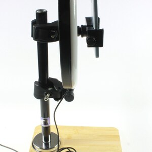 May include: A black and silver phone stand with a round white light attached to it. The stand is on a wooden base and has a black cord.