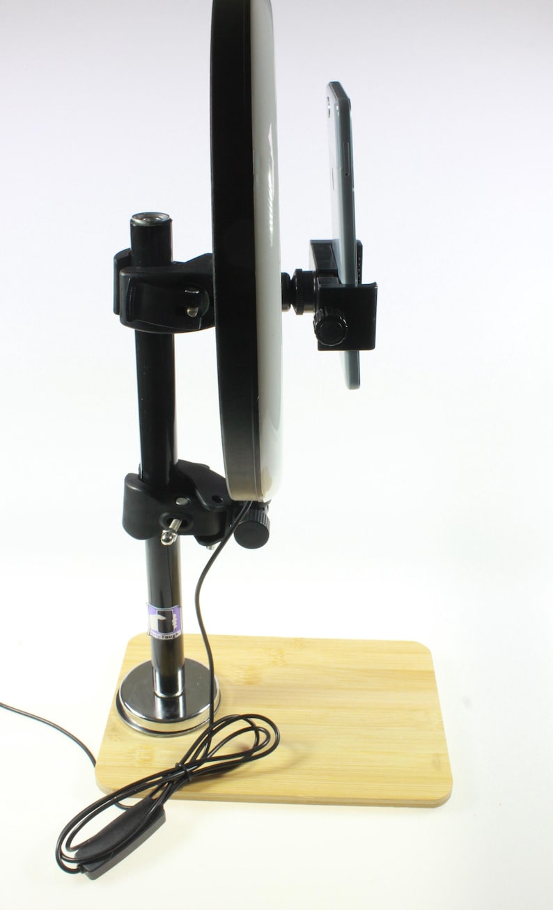 May include: A black and silver phone stand with a round white light attached to it. The stand is on a wooden base and has a black cord.