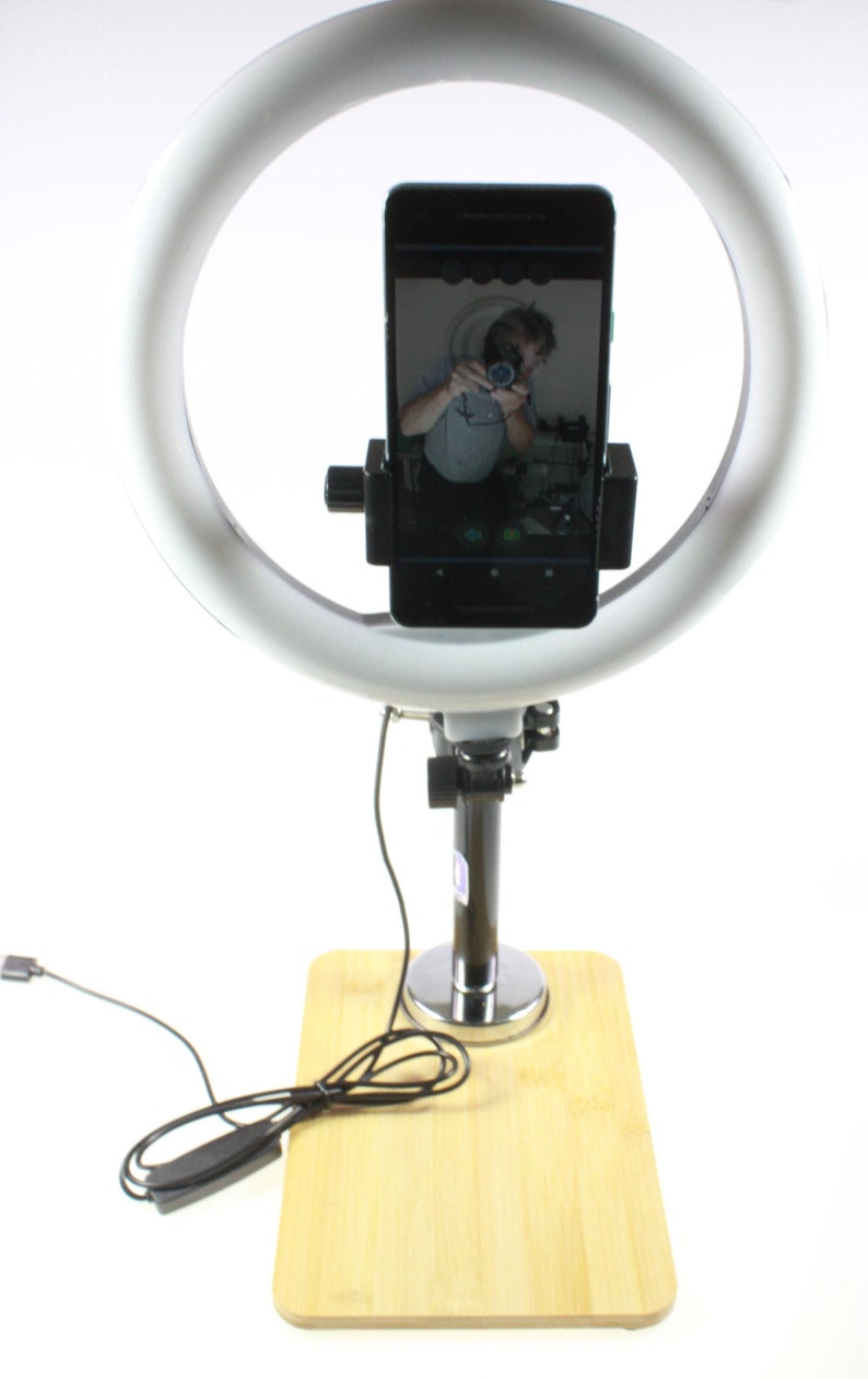 May include: A white ring light with a black phone holder and a black phone inside. The phone is taking a selfie of a person. The ring light is attached to a black stand with a silver base. The stand is on a wooden base.