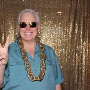 May include: A man wearing sunglasses and a blue shirt with a large gold chain necklace. He is making a peace sign with his right hand.
