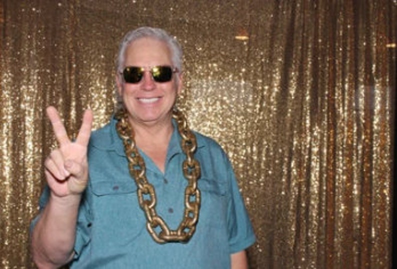 May include: A man wearing sunglasses and a blue shirt with a large gold chain necklace. He is making a peace sign with his right hand.