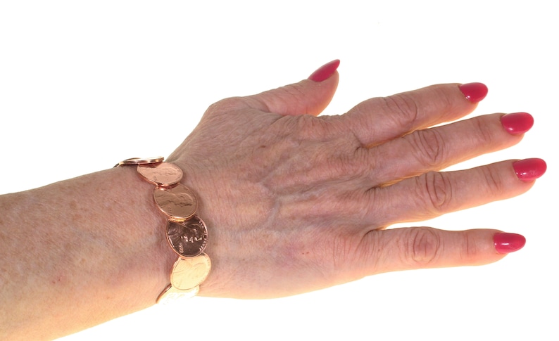 Peut inclure: Un bracelet en cuivre compos&eacute; de pi&egrave;ces de monnaie. Le bracelet est fait de pi&egrave;ces de monnaie li&eacute;es, affichant les faces et les piles des pi&egrave;ces. Le bracelet est port&eacute; au poignet, avec la main et les doigts visibles. Les ongles sont peints en rose vif.